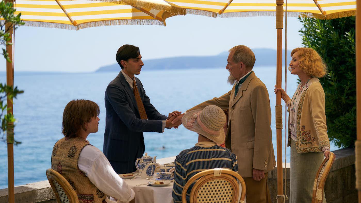 Virat and George shake hands under umbrellas in front of the sea, with Bella, Lucian, and Amelia watching