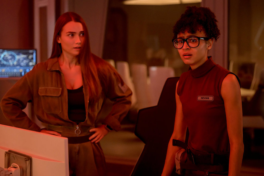 Eva and Alicia stand on the bridge of a ship in space with a red light.