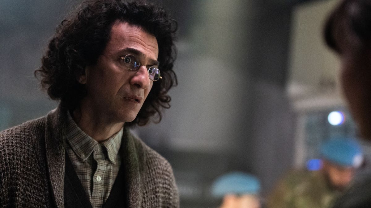 Dr. Nima Rousseau leans in on a conversation with Alex. He wears a tweed blazer over a dark vest and light green shirt buttoned up to the collar. 